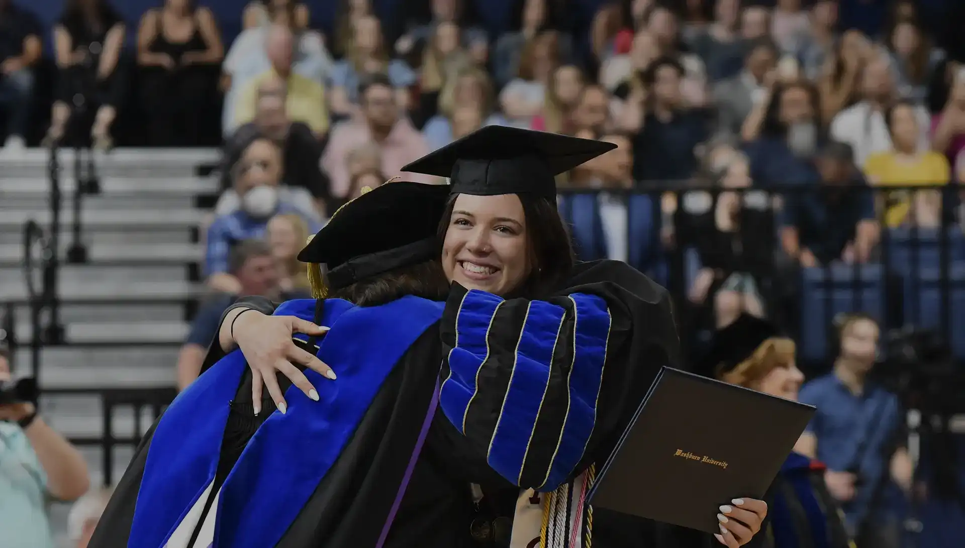 Student holding diploma hugging faculty member during commencement.