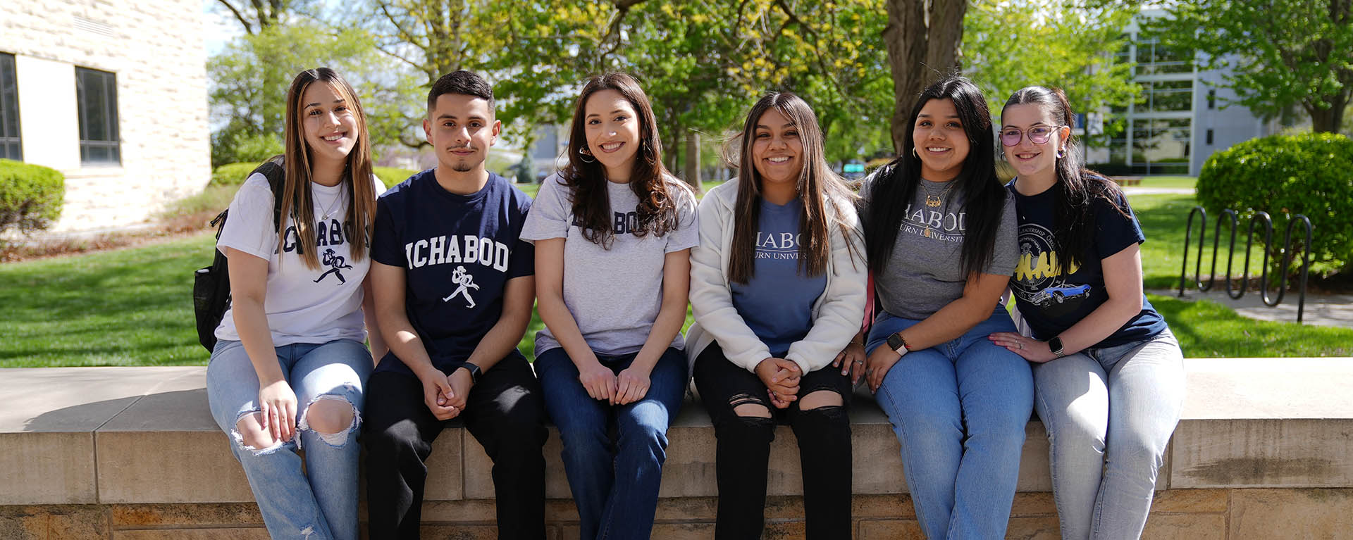 Students pose for a photo and smile while sitting on a limestone wall on campus.