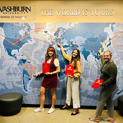3 students standing in front of a wall world map and pointing at locations
