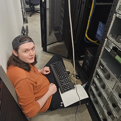 A computer information sciences student works in a server room with a keyboard on his lap.