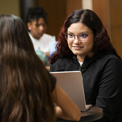 A student smiles at a friend while working on a laptop.