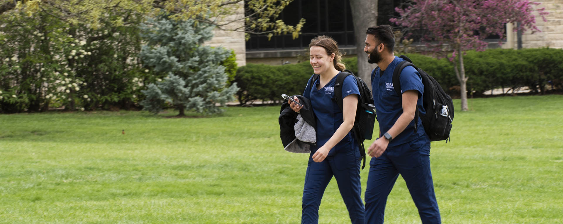 Two nursing students smile while walking across campus in their scrubs.